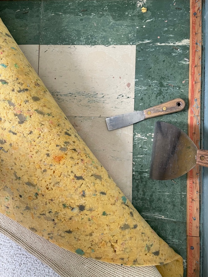 carpet tack strips in a corner with green and white linoleum, folded back yellow foam carpet pad and two putty knives