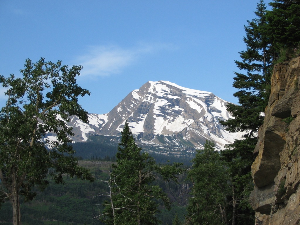 snow covered mountain in the distance against a blue sky with trees and craggy rock in the foreground