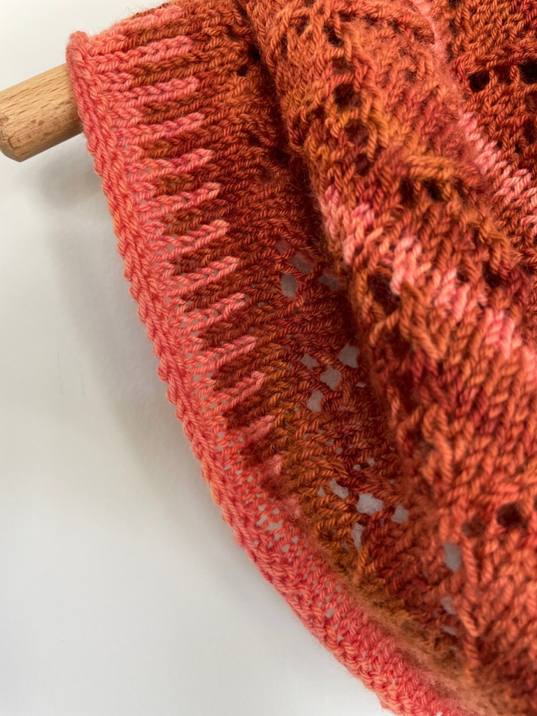 stitch detail of a handknit cowl in rust colored tones with a lace pattern of repeated diamonds on a wooden hanger