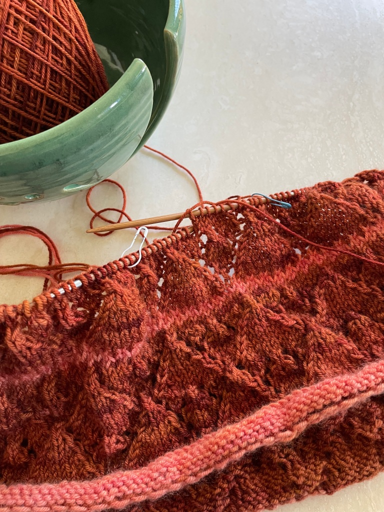 an incomplete handknit cowl in rust colored tones with a lace pattern of repeated diamonds resting on a marble background and a green yarn bowl in the upper left corner